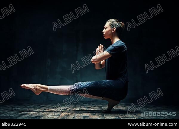 Young beautiful woman doing yoga asana tiptoe pose prapadasana variation on dark room. Practicing yoga; wellbeing; wellness concept.