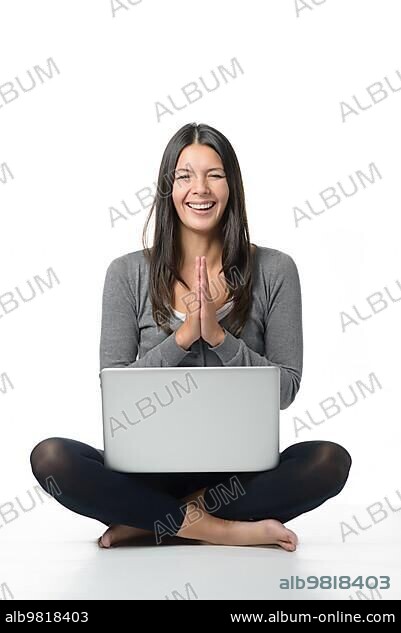 Happy young woman meditating with her laptop sitting cross legged and barefoot on the floor with her hands clasped as though in prayer and a beaming smile of joy; isolated on white.