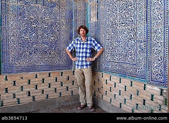 Young man at fine decorated wall with blue and white glazed tiles and zoroastrian symbols of ease Tash Hauli Palace Khiva Uzbekistan.