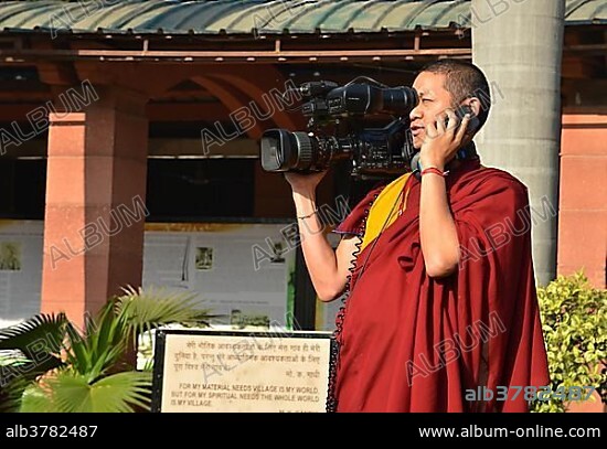 Tibetan monk in a red robe working as cameraman carrying a large video camera and a mobile phone, Global Buddhist Congregation 2011, at Smitri Gandhi, New Delhi, India, Asia.
