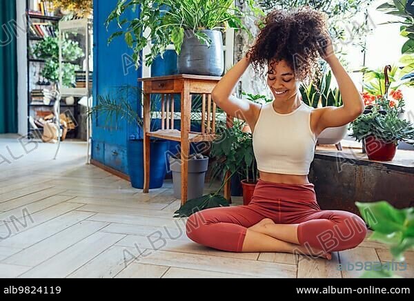 Young adult happy fit slim healthy African American ethnic woman wearing sportswear sitting in yoga pose at home in living room; doing fitness morning workout exercises training.