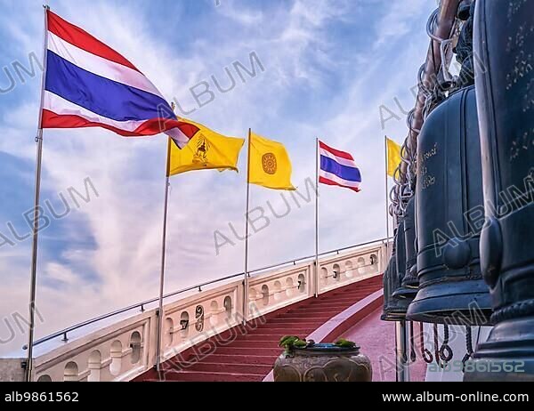 Row of Thai national, buddhism and Thai Royal flags along spiral staircase of red steps and white parapet, bells in blurred foreground. Stairway at Golden Mount temple or Wat Saket, Bangkok, Thailand. Incomplete inscriptions in Thai not translable.