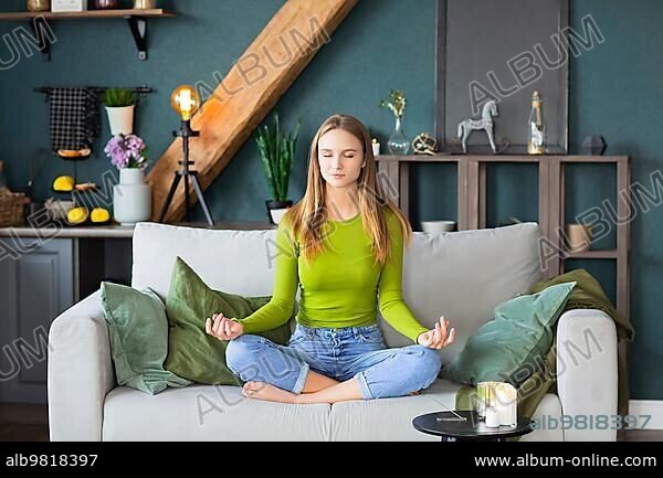Relaxed peaceful millennial blond girl with closed eyes sitting in lotus pose on couch in living room at home; taking break for meditation yoga practice. Stress relief and mental health concept.