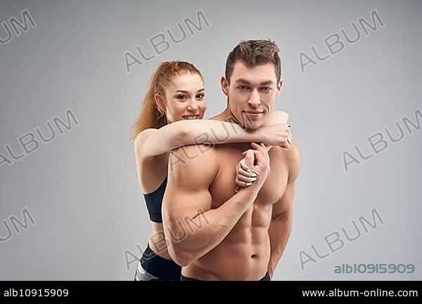 Side view of muscular shirtless man and reheaded woman with ponytail in sportswear hugging man from behind against grey background.