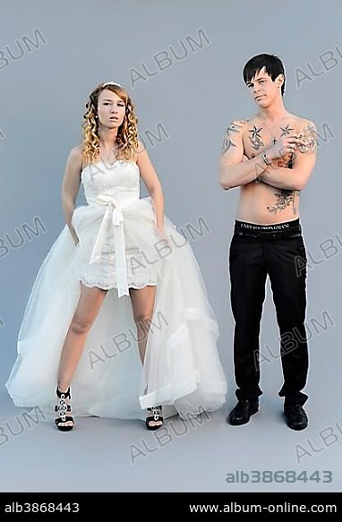 Wedding picture, a bride in a short dress and a bare chested groom with tattoos, holding a cigarette.