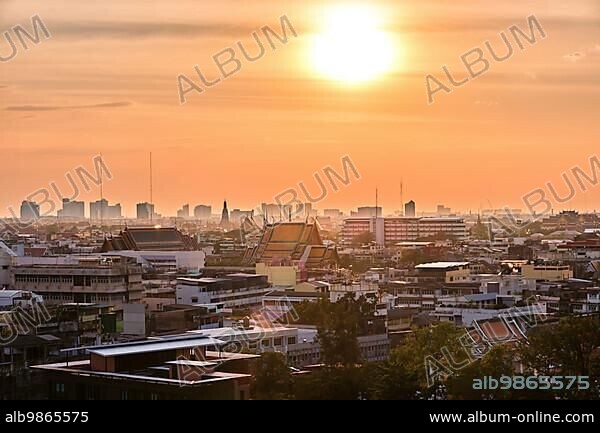 Beautiful cityscape of Bangkok city center in low sun, hazy orange sky and clouds, Thailand. Residential low-rise area in foreground, traditional temples, high-rises in downtown. Heat, summer, haze, heatwave, Asian climate, Buddhist religion.