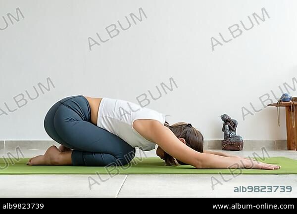 Beautiful woman practicing Balasana yoga asana; child's pose in yoga studio with Buddha statuette.