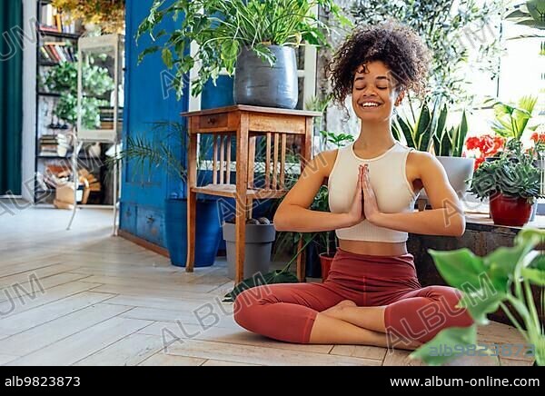 Young adult happy fit slim healthy African American ethnic woman wearing sportswear sitting in yoga pose at home in living room; doing fitness morning workout exercises training.