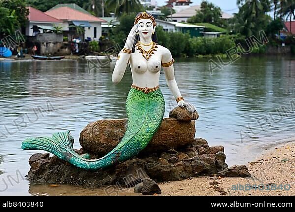 Greeting mermaid statue with white upper body and green tail fin sits on a stone on the shore, Buddhist temple Wat Phra Yai, Ko Phan Island, Koh Samui, Thailand, Asia.