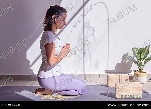 Woman meditating on her yoga mat with her hands in Anjali Mudra position in a peaceful setting with a Buddha statuette.