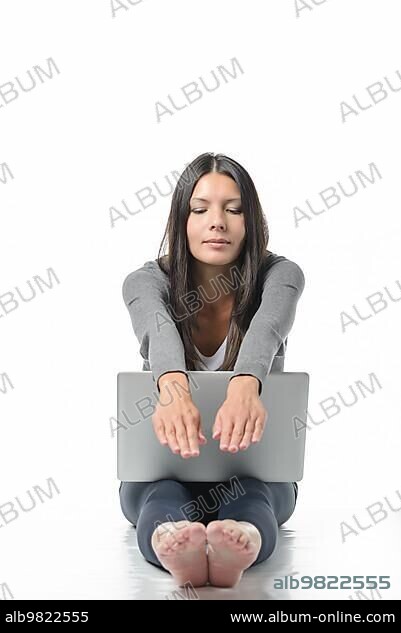 Attractive young woman sitting barefoot on the floor with her laptop computer stretching her hands and feet relaxing while meditating with closed eyes and a look of serenity in a wellness concept.
