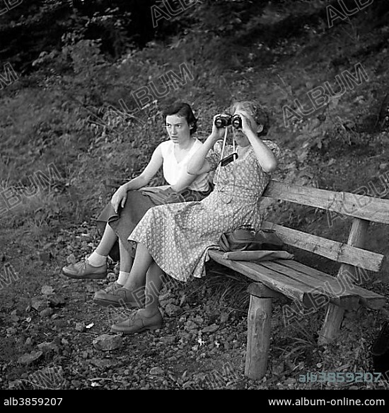 Two women are sitting on a bench and one of them is looking through binoculars, ca. 1945 to 1955, near Freiburg, Germany, Europe.