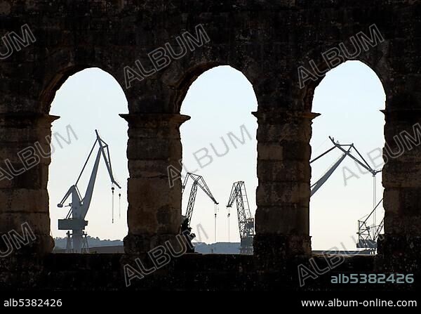 Harbour cranes seen through arches of ancient Roman amphitheater, arena, in Pula, Istria, Croatia, Europe.