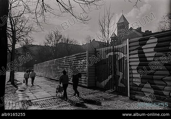 GDR, Berlin, 22. 3. 1988, Schönhauser Allee artistically designed building fence . . and Sacred Heart, Jesus, church in the background, couple with pram.