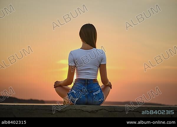 Rear view of a woman in meditation pose on the pier during summer sunset.