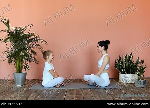 Side view of smiling woman and little girl practicing yoga pose while sting on mat in cozy living room and looking away.