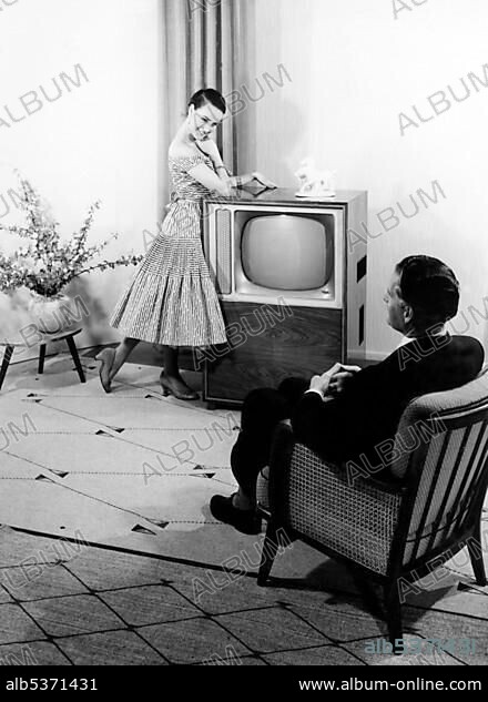 Woman posing in front of a TV set, man sitting in an armchair, historical photo, circa 1955.