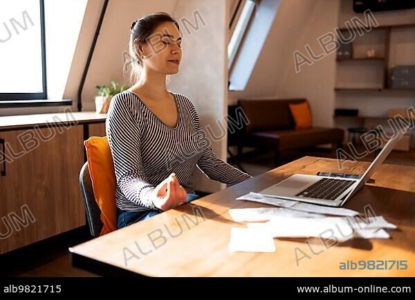 Stress relief during freelance work. Brunette woman freelancer sitting in lotus pose and meditating with hands in mudra gesture on chair in front of open laptop on table; working from home.