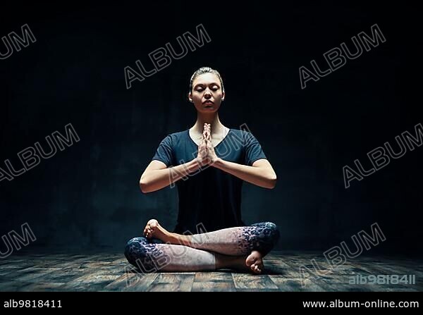 Young sporty woman practicing yoga doing Ardha Padmasana exercise with namaste in dark room. Lotus pose; meditation concept.