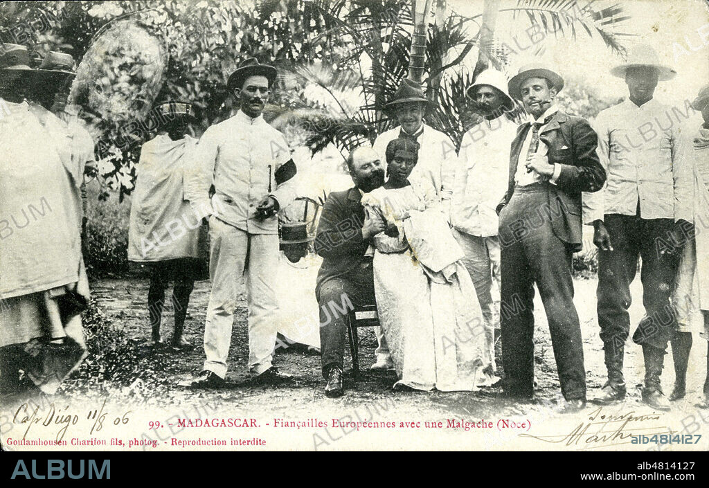 5 hommes blancs assez goguenards, entourant une femme malgache assise sur les genoux de l'un d'eux : 'Madagascar. Fiancailles europeennes avec une malgache (noce)'. Une ambiance etrange se degage de cette image, on a du mal a croire que ce n'est pas une situation abusive. Carte postale de 1906. Credit : Collection IM/KHARBINE-TAPABOR.