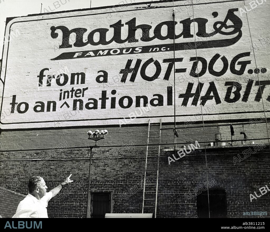 Entitled: "From a hot dog to an international habit" showing Nathan Handwerker pointing to restaurant sign painted on outside wall.  Nathan's Famous, Inc. is an American company that operates a chain of fast food restaurants specializing in hot dogs. Nathan's began as a nickel hot dog stand in Coney Island in 1916 and bears the name of co-founder Nathan Handwerker who started the business with his wife, Ida, who created the hot dog recipe they used, and Ida's grandmother created the secret spice recipe. The original Nathan's still exists on the same site that it did in 1916. Having been open for business every day, 365 days a year, the stand was forced to close on October 29, 2012, due to Hurricane Sandy. Service is provided year round inside, and during the summer additional walk-up windows are opened to serve the larger seasonal crowds. orld Telegram & Sun photo by Roger Higgins July 22, 1959.
