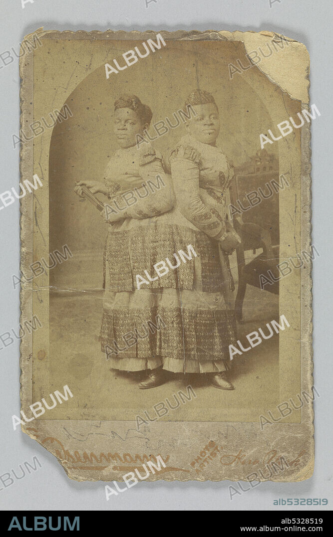CHARLES EISENMANN. A photographic print of African-American conjoined twins whose stage names were "The Carolina Twins", "The Two-Headed Nightingale" and "The Eighth Wonder of the World". The photograph features the women standing facing the camera. They are wearing a long-sleeved dress with two bodices. A chair is partially visible in the back right of the photograph. The photograph is mounted on heavier paper that has a scalloped edging and the name of the photography studio printed in gold at the bottom. The back of the photograph has a partial inscription of name.