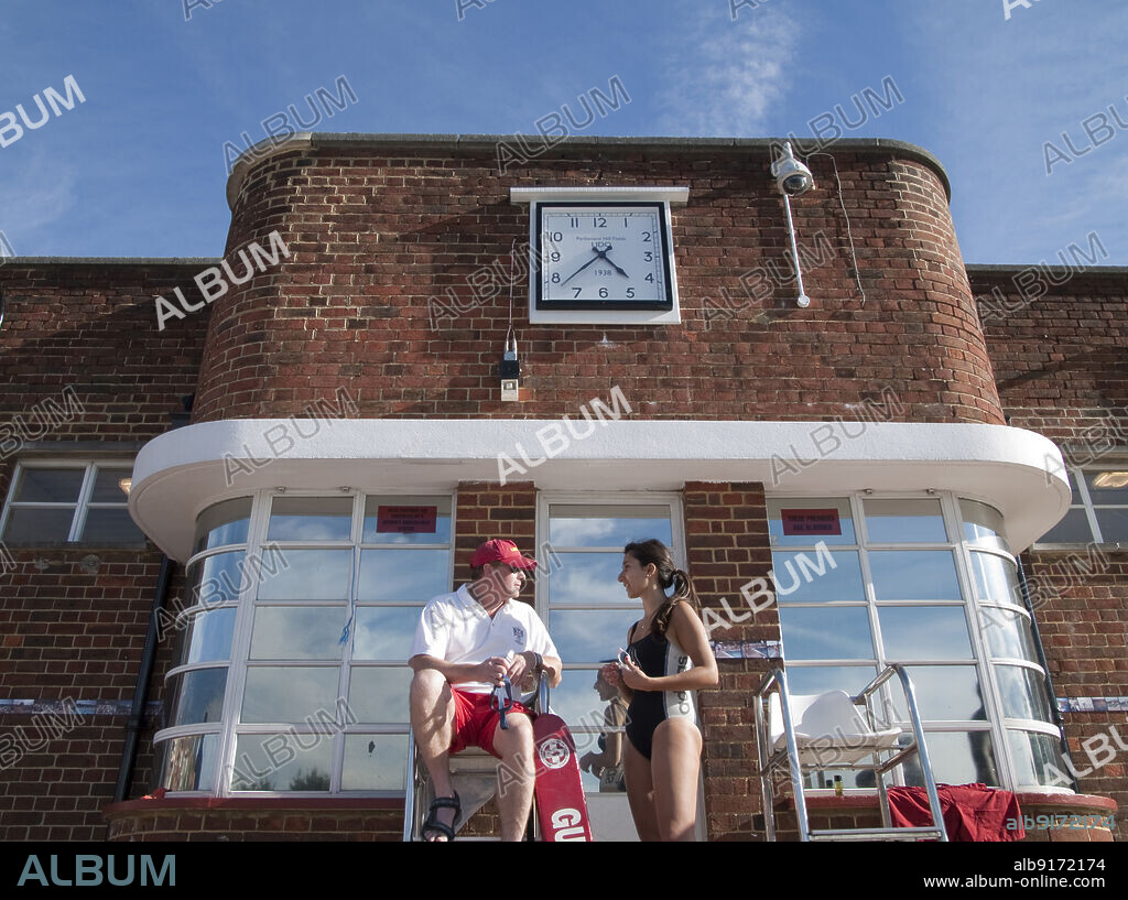 SIMON INGLIS. Parliament Hill Fields Lido, Gordon House Road, Gospel Oak, Camden, Greater London Authority, 2012. A lifeguard and swimmer talking beneath the square clock on the central admin office at Parliament Hill Fields Lido in Gospel Oak. Parliament Hill Fields Lido opened in 1938 and was designed by Harry Arnold Rowbotham and T L Smithson. It was one of thirteen lidos built by London County Council between 1909 and 1939. The pool remained open until major refurbishment in 2004-2005. Original features, such as the main clock, signage, and aerator fountain, have been restored or replicated. It is Grade II listed.