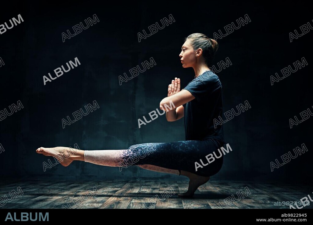 Young beautiful woman doing yoga asana tiptoe pose prapadasana variation on dark room. Practicing yoga, wellbeing, wellness concept