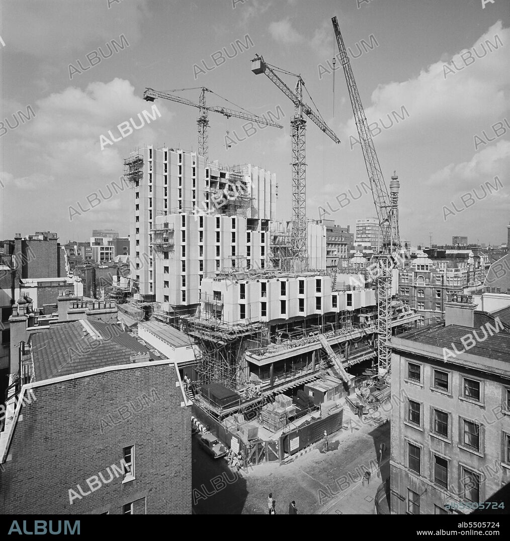 JOHN LAING PLC. The construction of the eastern three tower blocks of the Central London YMCA, showing cranes atop and alongside the structure, viewed over rooftops from the east. It was announced in February 1971 that Laing had started work on a new building to replace an existing building which had opened in the 1910s. The project comprised four residential towers between 6 and 12 storeys high, above a podium and basements. The towers would accommodate 1,200 residents in over 700 single and double bedrooms; staff would be accommodated in penthouse suits on the top of the three lower towers. The towers were built on a two-storey podium containing restaurants, offices, a public library, and shops. Beneath the buildings were basements 16 metres below street level, for car parks, a swimming pool, a gymnasium, a sports hall, and a conference centre. The basements and podium, which were built using reinforced concrete and structural steel, comprised Phase I of the project; the podium was built onto the foundation piles before the basements had been excavated. Construction of the towers began earlier than originally planned: excavated material was removed from the basements using conveyors rather than mounting cranes onto the podium frame, on which the towers were to be built. The towers were clad in 2,000 precast concrete panels and had deeply moulded white concrete window panels, both of which were manufactured by John Laing Concrete in Princes Risborough. The towers were built in Phase II and the building was completed in 1977. A cropped version of this photograph was published in the January 1975 edition of 'Team Spirit', Laing's newsletter.