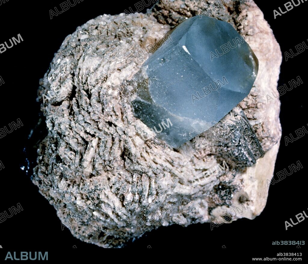 Topaz. View of a blue topaz in celandite. Topaz is a silicate mineral made of aluminium silicate. Topaz is a very hard stone, often transparent or translucent with a vitreous lustre. It is often colourless, but can be coloured, yellow, blue (as here), pink, green, violet or reddish-yellow. Clear or finely coloured varieties have always been used as gemstones. This specimen was found in the Ural Mountains, Russia.
