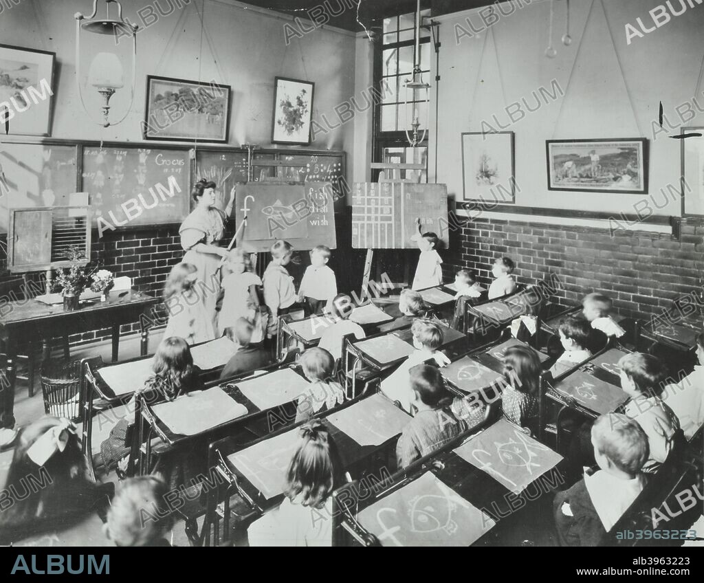Spelling lesson, Southfields Infants' School, Wandsworth, London, 1907.  Children are being taught the formation of the word 'fish' by a teacher. The children all have pictures they have drawn of fish on their desks in front of them. A boy at the front of the class has his hand raised to ask a question.