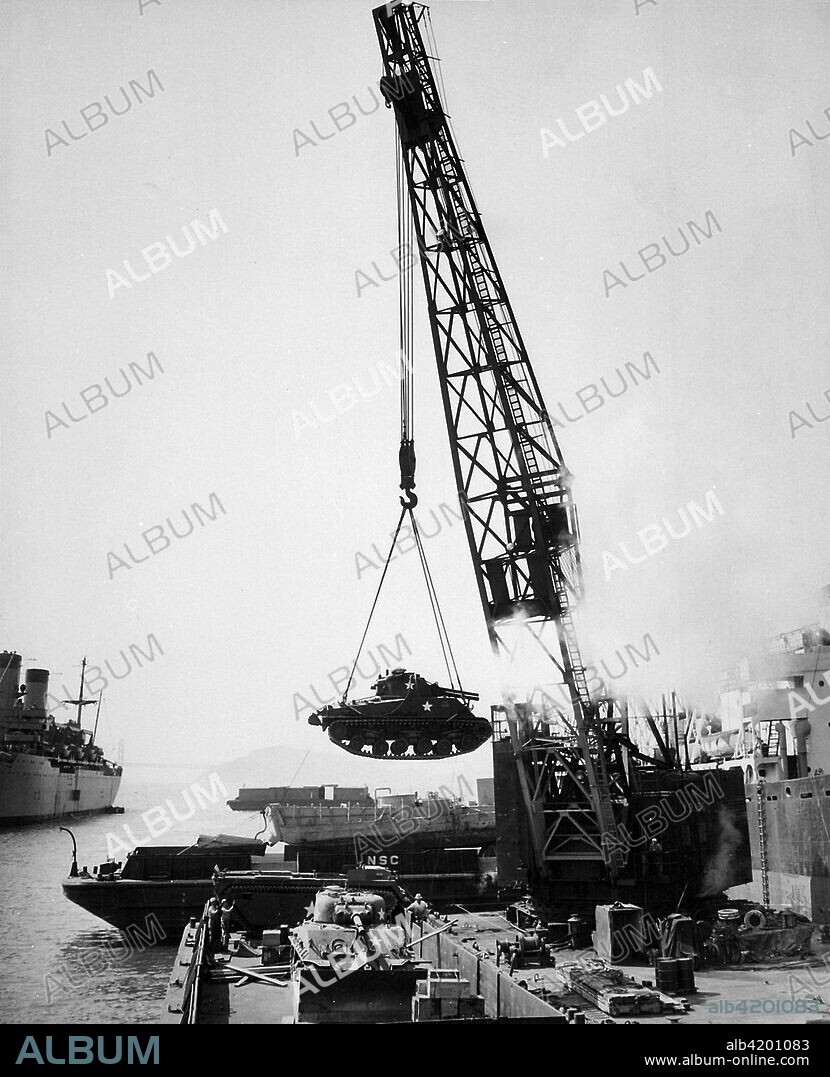 A U.S. Marine Corps M4 Sherman tank is swung aboard a barge at the Naval Supply Center at Oakland, California (USA) by crane, for transshipment to Korea, 1950. Another M4 and an LVT are visible on the barge.