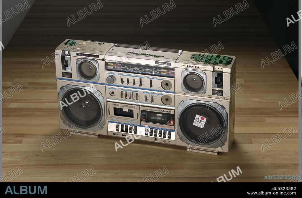 A tecsonic boombox with a light brown metal cover used by Public Enemy. The boombox has a handle at the top, collapsible antenna, six speakers, a radio, a double cassette player, and a number of controls. There are green and black Public Enemy stickers adhered around the left, top, and right sides. There is one sticker on each side and three on the top. All of the stickers have varying degrees of loss. Two cassette compartments marked "METAL / 4 BAND STEREO RECEIVER / DOUBLE CASSETTE PLAYER" and "5-BAND GRAPHIC EQUALIZER MX-900." The large round speaker at the bottom right has a white sticker with black and red print that says "I (heart symbol) / SOWETO / www.iluvsa.co.za / Where: CHAF - POZI / When: 3rd Saturday / of every month." There is a black label with silver text on back of speaker. It reads "TECSONIC / STEREO DOUBLE CASSETTE RECORDER / WITH 4-BAND RADIO / MODEL NO: MX-900 / AC 110/220V 50/60Hz30W / DC 15V"D"SIZE BATTERY x10PCS / MADE IN KOREA.".