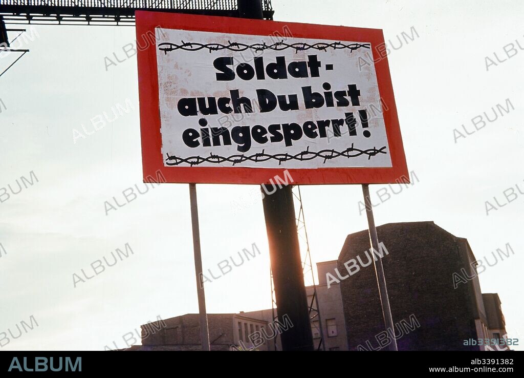 Red and white sign with barbed wire graphic on the border between West Berlin and East Berlin, during the Cold War, with text in German reading "Soldat auch du bist eingesperrt" or "Soldiers you are also imprisoned", encouraging East German soldiers to defect to the West, Germany, 1975.