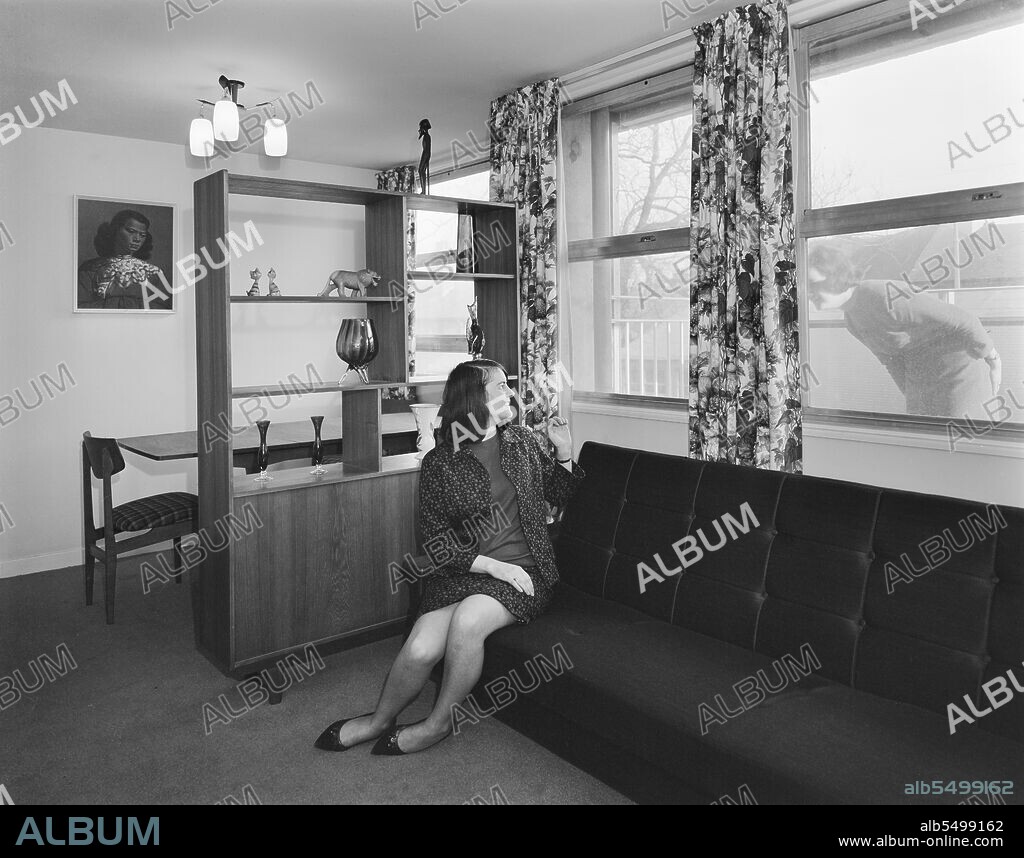 JOHN LAING PLC. Bearwood House, Bearwood Road, Smethwick, Sandwell, 02/02/1966. A young woman sitting in the lounge-diner of a flat at Bearwood House, waving to a friend outside. 'Sectra' was a French prefabricated steel formwork design for flats which John Laing and Son Ltd acquired the British rights to in 1962. It was a method of using precision made steel formwork for the placing of structural concrete in 'tunnel' sections in room unit widths and ceiling heights. The units were bolted together in rows on special tracks, with the concrete poured to form the walls and floors in one operation. The formwork was internally heated to accelerate the hardening of the concrete in the mould and the sections were then lifted into position by a tower crane on the construction site. Bearwood House was built by Laing for Smethwick Borough County Council. The architects were K.R. Emmett, S. Greenwood, J.E. Hepburn and R.E. Philcock. Construction work started in 1965 and the building was opened on 25th January 1966. It has since been demolished.