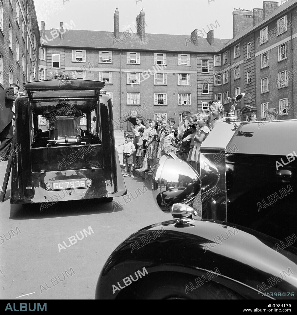 Funeral on a council estate, London, 1960-1965. A hearse parked in the courtyard of a tenement block, the bonnet of another car in the foreground partly obscuring a line of children standing watching, in either Islington or Camden.