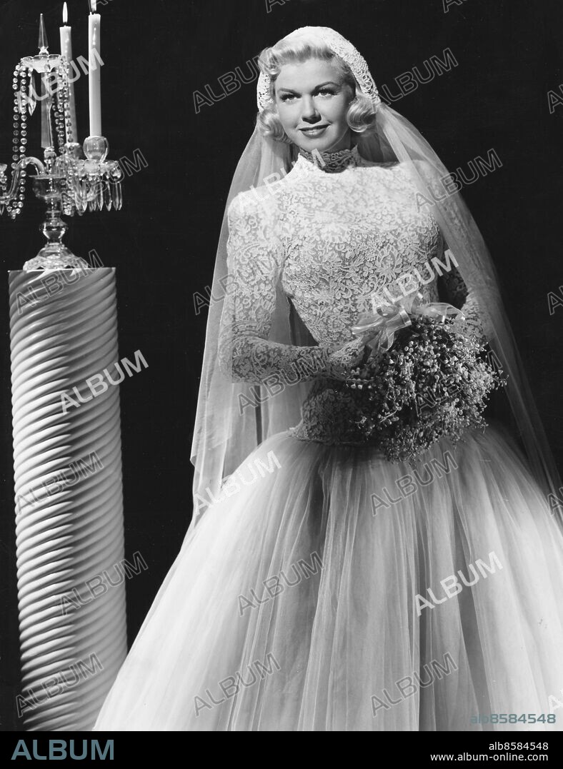 Orig. bildtext... FOR YOUR WOMAN'S PAGE Hollywood, Calif. Molding white imported lace over natural souffle makes a lovely wedding gown modeled by Doris Day, Warner Bros. star. Shaped to the body's lines the long-sleeved, high-throated bodice tops a flowing skirt of tulle and beneath it all a whispering slip of white tarfeta. For the bridal illusion, a sweep of tulle is fastened to the caplet (an heirloom) of imported white lace that fits snugly to the head. The bridal bouquet in this Milo Andersson creation is a ball of lillies of the valley. Anm. Skådespelare Liljekonvaljer Buketter Ljusstakar Spets Kandelabrar persons: Doris Day sites: USA.