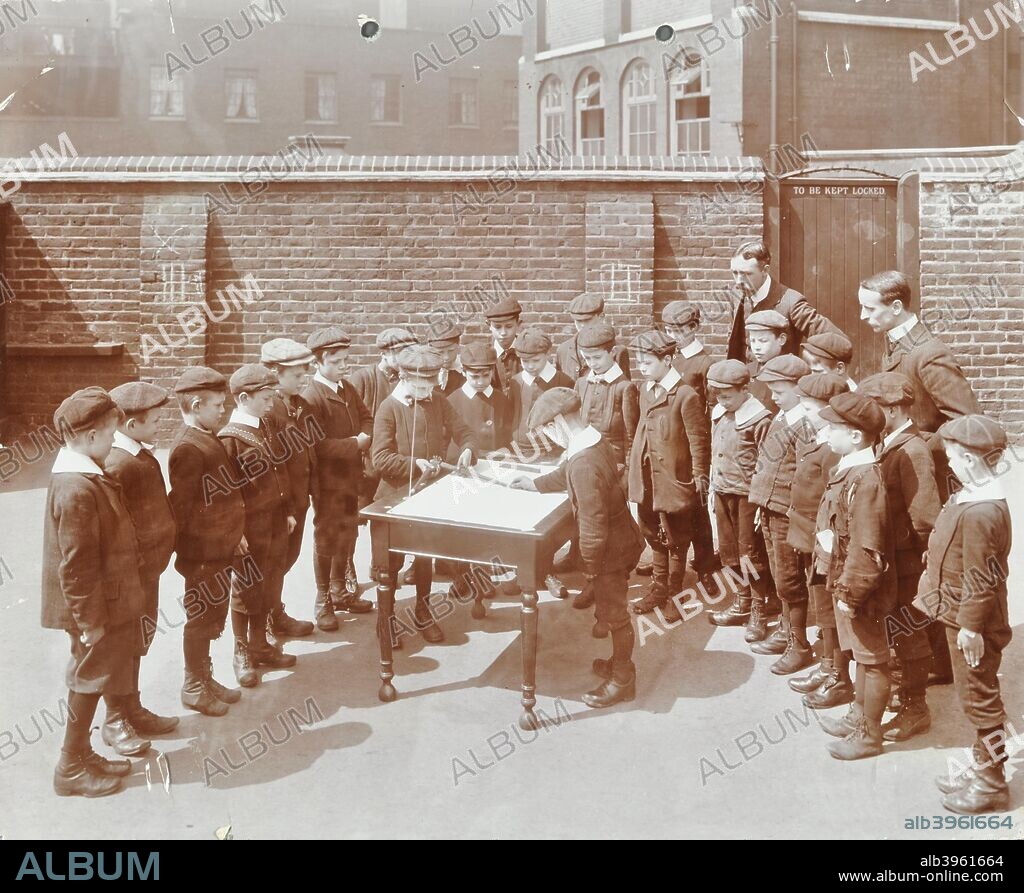 Geography lesson, Hague Street School, Bethnal Green, London, 1908. Two boys in the playground trying to find true north, watched closely by their classmates and teachers.