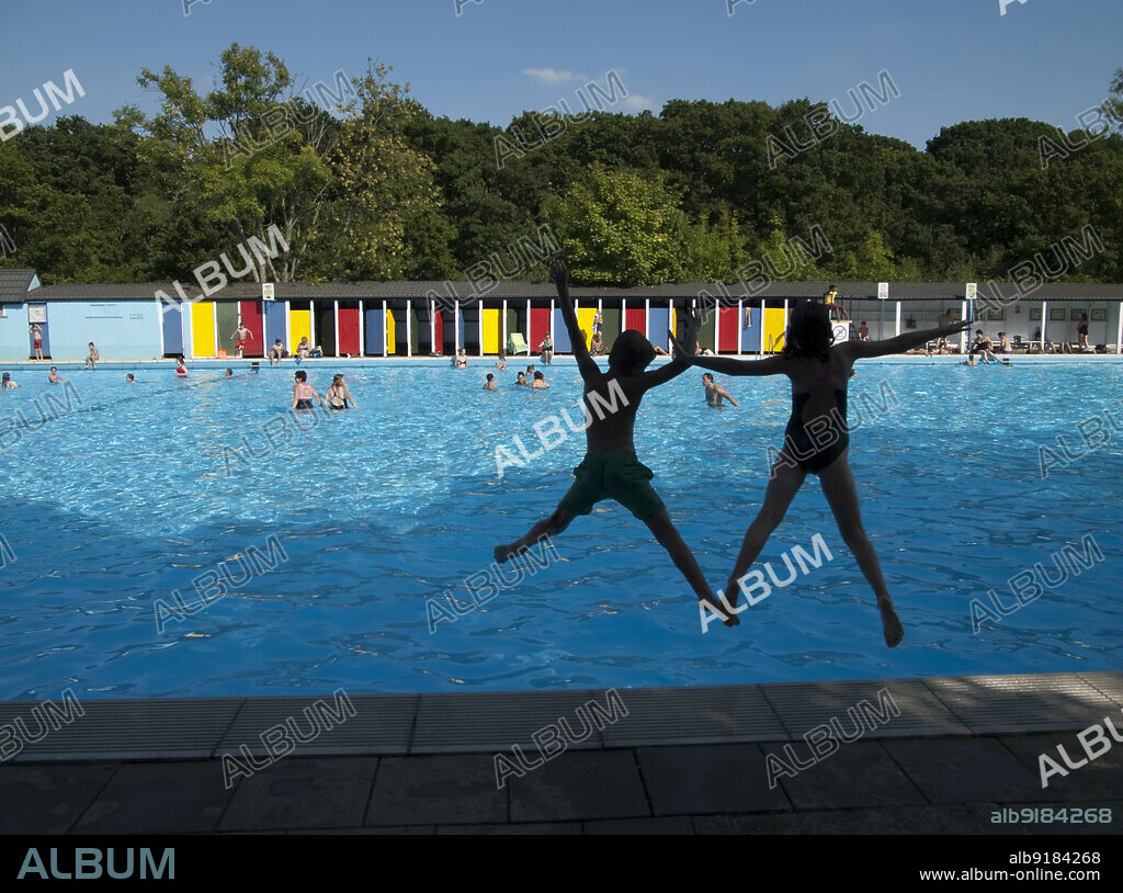 SIMON INGLIS. Tooting Bec Lido, Tooting Bec Road, Streatham, Wandsworth, Greater London Authority, 2013. Two children, silhouetted against the water, jumping into the pool at Tooting Bec Lido, with colourful cubicles on the east side of the pool beyond. Tooting Bec Lido is London's largest and oldest outdoor pool. It was opened in 1906 and was originally known as Tooting Bec Bathing Lake. The pool measures 300 ft by 100 ft, making it twice the length of an Olympic pool and wider than a standard 25m pool is long. It holds one million gallons, and 1,400 swimmers can be in the water at any one time. A cafe was built in 1936. Renovation to the pool and cafe took place in 1999-2002, with a new paddling pool and entrance block were built, designed by WM Architects.