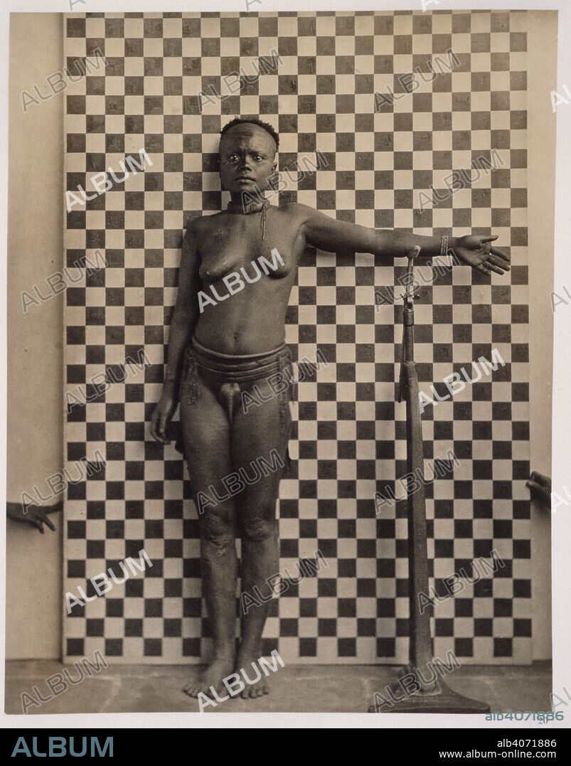 MAURICE VIDAL PORTMAN. Woman of the TÃ¡-KÃ©da tribe. Andamese Islanders, Volume 11. 1893. A full length study of a woman from the TÃ¡-KÃ©da tribe, aged about 45 years. She is posed in front of a checked screen of 2 inch squares (to indicate scale). The left hand is fully extended horizontally and rests on a stand. Image taken from Andamese Islanders, Volume 11. Originally published/produced in 1893. Source: Photo 188/11(20),.