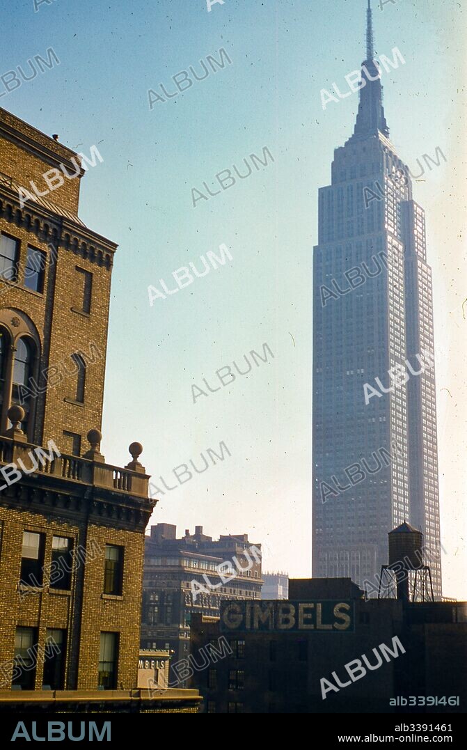 View facing northeast of the Empire State Building at 34th Street and Fifth Avenue in Manhattan, New York City, 1957. In the foreground a warehouse building with water tower stands in the shadows, with a painted advertising for Gimbels Department Store. In center frame, the corner edge of the Gimbels Store roof located at Sixth Avenue and 32nd Street is visible.