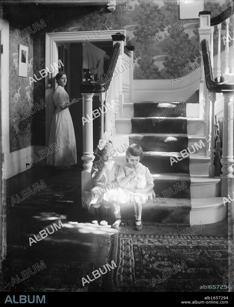 Lollipops': Posed photograph of two small girls sitting at the bottom of the stairs in a shaft of sunlight, sucking lollipops, while a woman watches from a doorway. Waban, Mass. Children in domestic interior, c1910.