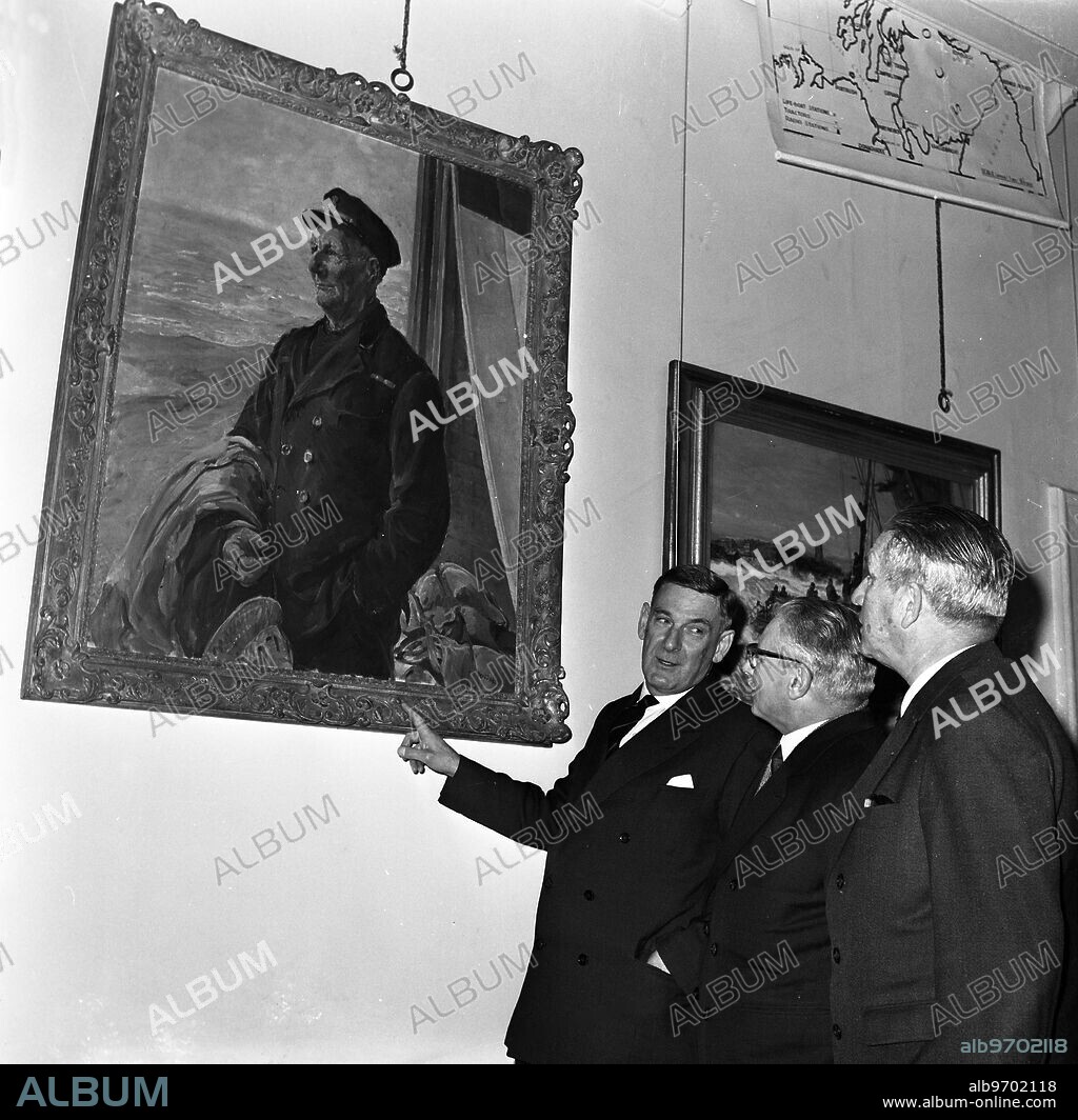 Britain's Chief Inspector of Lifeboats Commander Dutton (left) , describes a painting of Britain 's most famour lifeboat coxswain Henry Blogg , G.C. , BEM to Captain Rogerio Pinto ( centre) and Captain Carlos Carre Ira (right) , members of the Portuguese Lifeboat Society , at the Royal National Lifeboat Institution 's London headquarters . The Portuguese lifeboat officials are on a 5-day visit to the United Kingdom to visit RNLI establishments and the Board of Trade . 6 April 1970.