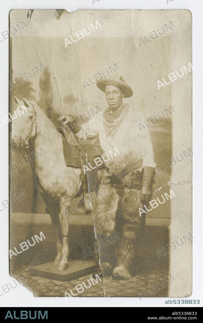 This real photo postcard depicts a man in a cowboy costume posing with a fake horse. He is standing to the right of the horse and is wearing a long-sleeve shirt, pants, fur chaps, a hat and a scarf around his neck. His right arm is resting on the horse's saddle. The backdrop behind him depicts a nature scene. The base of the fake horse and the floor of the photo studio are visible at the bottom of the image. The image is oriented in the bottom left corner of the postcard, leaving blank space at the top and on the right side. On the back of the postcard the word [POST CARD] is printed at the top. The words [CORRESPONDENCE HERE] and [NAME AND ADDRESS] divide the back into two sections. Text marks the spot for postage in the top right corner. The word [AZO] repeats forming a square with a triangle in each corner. The words [PLACE / STAMP / HERE] are printed inside the square. There are no inscriptions, front or back.