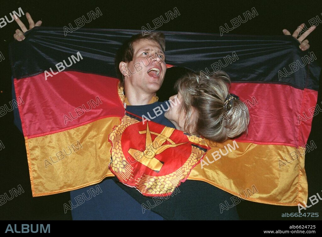 Two young people from East Berlin celebrate the immininent German reunification with a GDR flag in Berlin, former GDR, 02 October 1990. Almost one million people celebrated the German reunification at midnight. 45 years after the end of World War II, the German citizens are reunited again in a sovereign state. Prior to that, the regime of SED (Socialist Unity Party of Germany) had collapsed rapidly. Urged by the population, GDR opened its borders on 09 Nobember 1989. 02/10/1990
