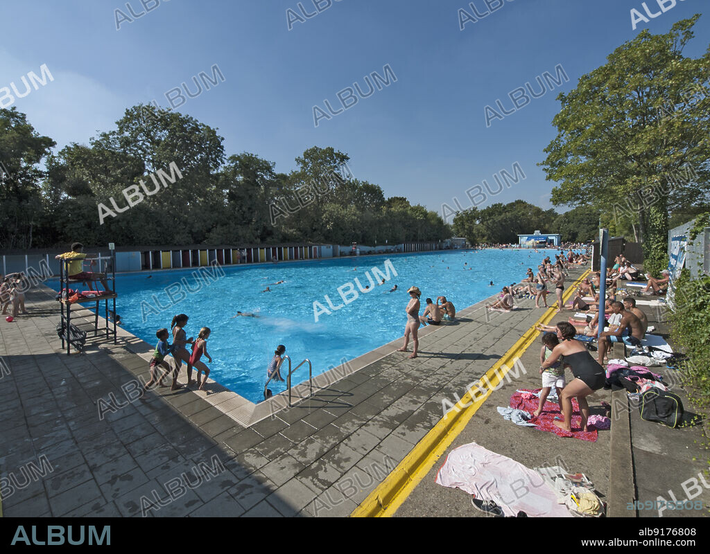 SIMON INGLIS. Tooting Bec Lido, Tooting Bec Road, Streatham, Wandsworth, Greater London Authority, 2013. An elevated view of the swimming pool at Tooting Bec Lido, Streatham, busy with swimmers and sunbathers, seen from the south-east corner. Tooting Bec Lido is London's largest and oldest outdoor pool. It was opened in 1906 and was originally known as Tooting Bec Bathing Lake. The pool measures 300 ft by 100 ft, making it twice the length of an Olympic pool and wider than a standard 25m pool is long. It holds one million gallons, and 1,400 swimmers can be in the water at any one time. A cafe was built in 1936. Renovation to the pool and cafe took place in 1999-2002 by WM Architects, and modern indoor facilities were added to the south end in around 2015, designed by David Gibson Architects.