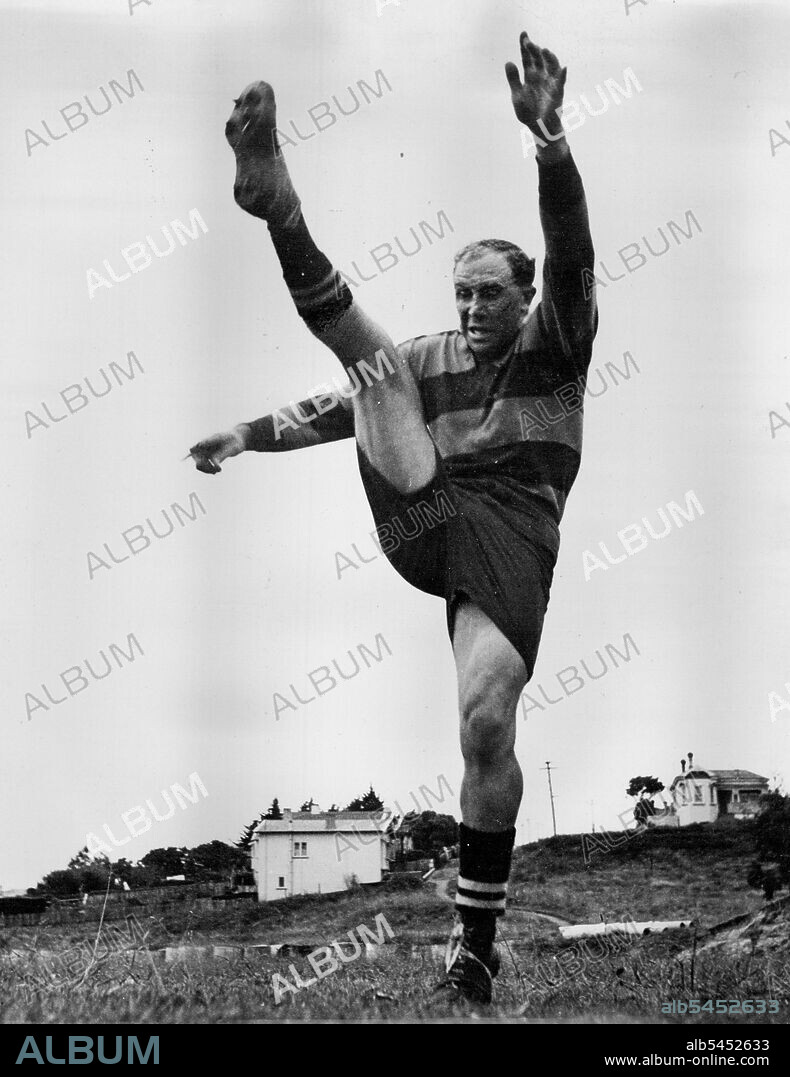 In the grey light of dawn Bob Scott limbers up his kicking leg in preparation for the approaching 1948 Rugby season in Auckland. Although he is very strongly built he is as supple as a ballet dancer/as this picture shows. He is wearing his Ponsonby Club jersey. July 13, 1951.