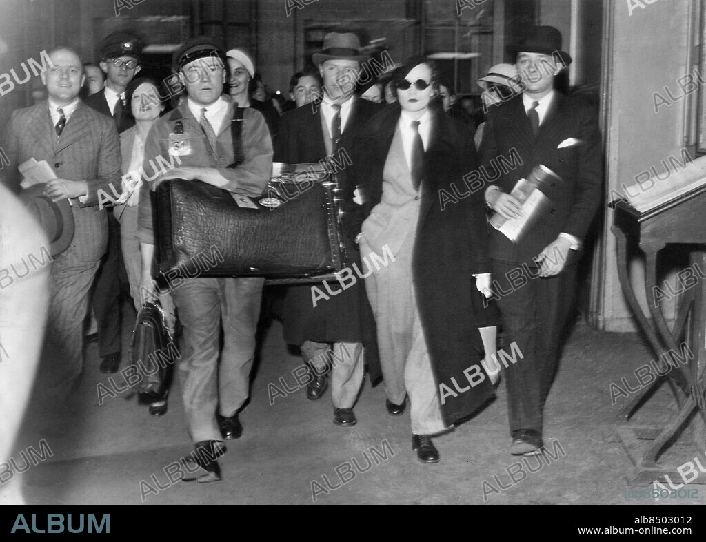 Orig. bildtext... TAKE 'EM OFF - WE KNOW YOU! Marlene Dietrich, arriving at the Gare St. Lazare, Paris, bound for a three months holiday in France, chose to continue her disguise of men's clothes and brown sunspectacles. On her right, her husband, Mr. Sieber; on her left, Marcel Boursier, who has charge of her French visit. Marlene's polo coat was a deep reddish brown, matching her beret. The suit she wore was of twill. May 19, 1933 Anm. M Dietrich, 1901-1992, tysk skådespelerska och sångerska, amerikansk medborgare 1939. R Sieber, 1897&2;1976, tjeckisk filmassistent, gift med M Dietrich till sin död. Bagage Basker Filmstjärnor Solglasögon CD696 persons: MARCEL BOURSIER;RUDOLF SIEBER;MARLENE DIETRICH sites: TYSKLAND*;PARIS;TJECKOSLOVAKIEN*;FRANKRIKE.