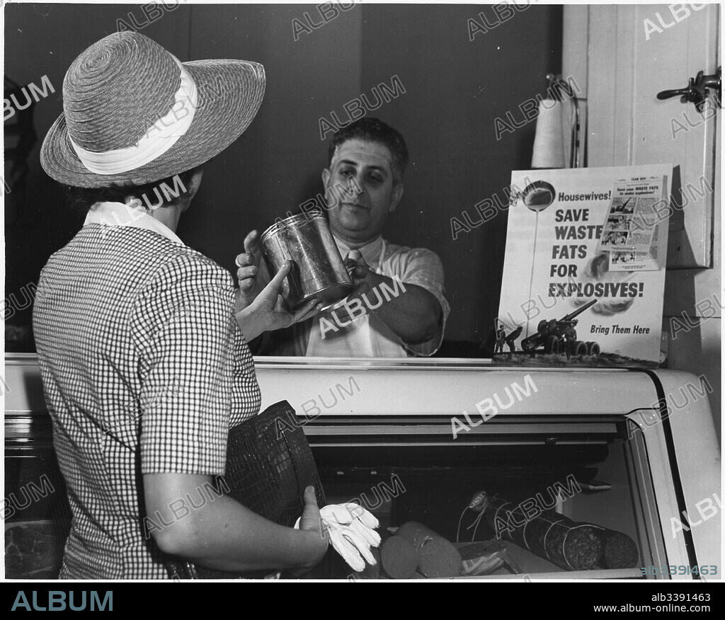 A housewife wearing a straw hat and white gloves reaches over the glass counter to hand a butcher a can of solid fat in exchange for money, after which it will be processed into ammunition for America's World War II efforts, 1942.