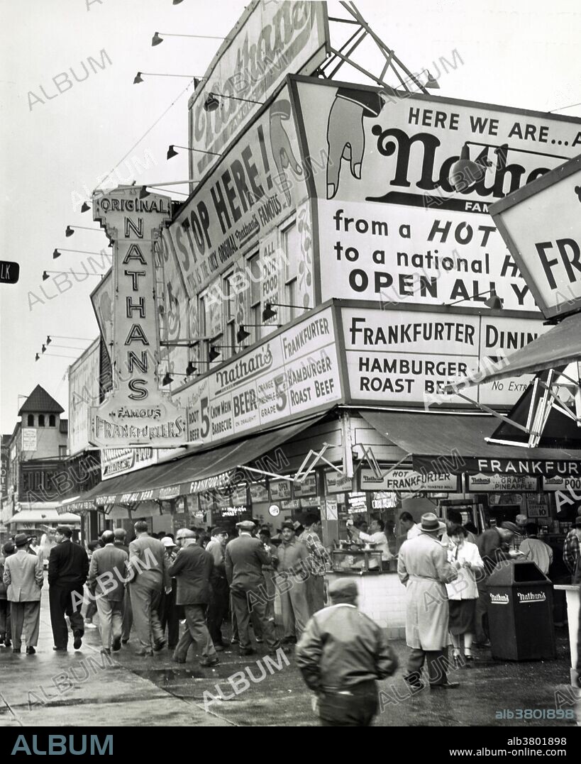 Entitled: "Nathan's Hot Dogs from boardwalk." Nathan's Famous, Inc. is an American company that operates a chain of fast food restaurants specializing in hot dogs. Nathan's began as a nickel hot dog stand in Coney Island in 1916 and bears the name of co-founder Nathan Handwerker who started the business with his wife, Ida, who created the hot dog recipe they used, and Ida's grandmother created the secret spice recipe. The original Nathan's still exists on the same site that it did in 1916. Having been open for business every day, 365 days a year, the stand was forced to close on October 29, 2012, due to Hurricane Sandy. Service is provided year round inside, and during the summer additional walk-up windows are opened to serve the larger seasonal crowds. World Telegram & Sun photo by Al Ravenna May 11, 1954.